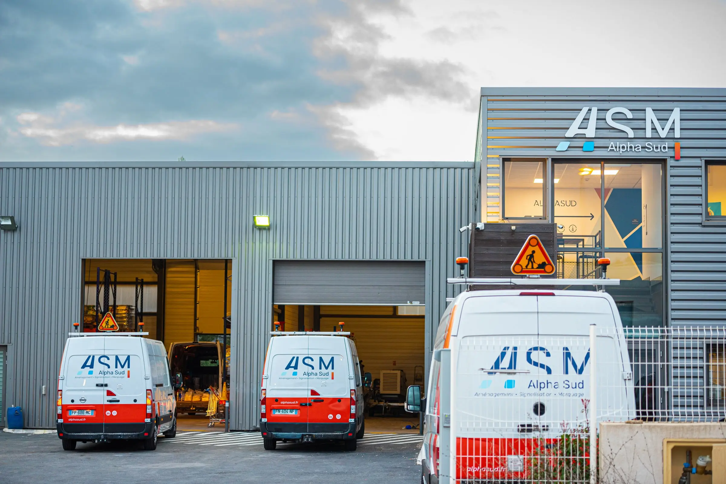 Les fourgons ASM Alpha Sud alignés devant l’atelier éclairé, au crépuscule