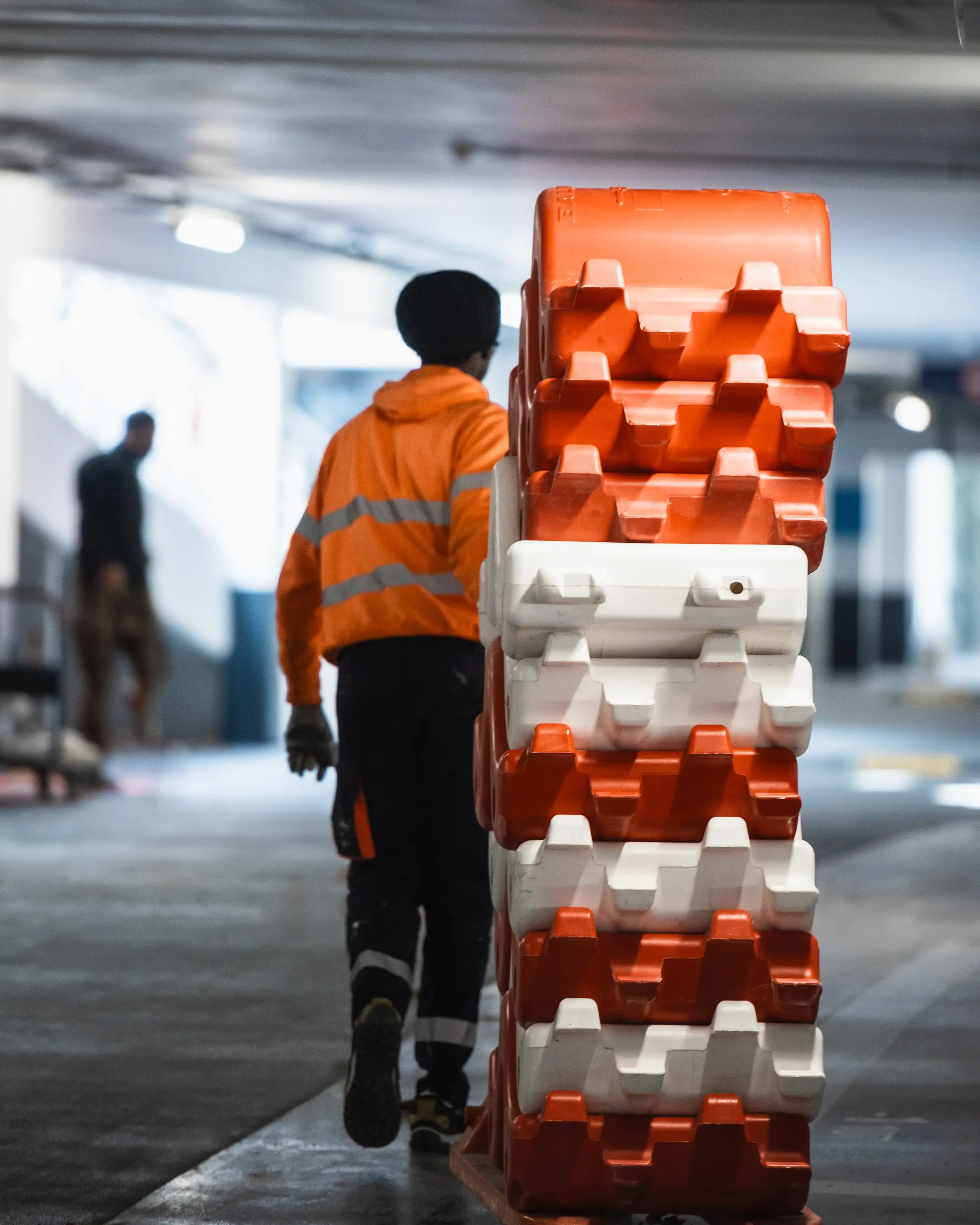 Salarié ASM transportant des barrières de chantier rouge et blanc empilées dans un parking