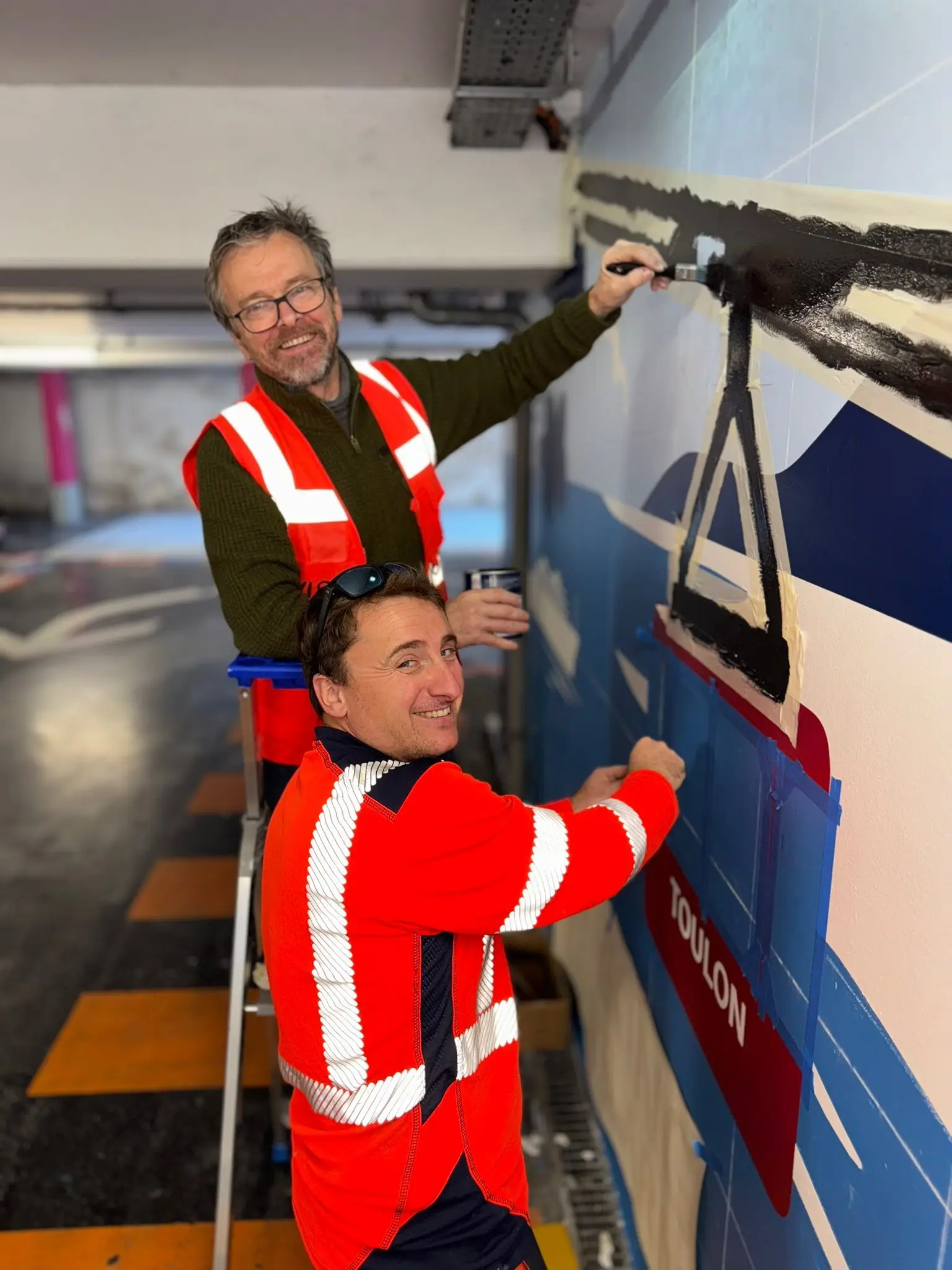 Deux salariés ASM peignant la fresque du parking de la gare de Toulon, silhouette de navire et lettrage « Toulon »