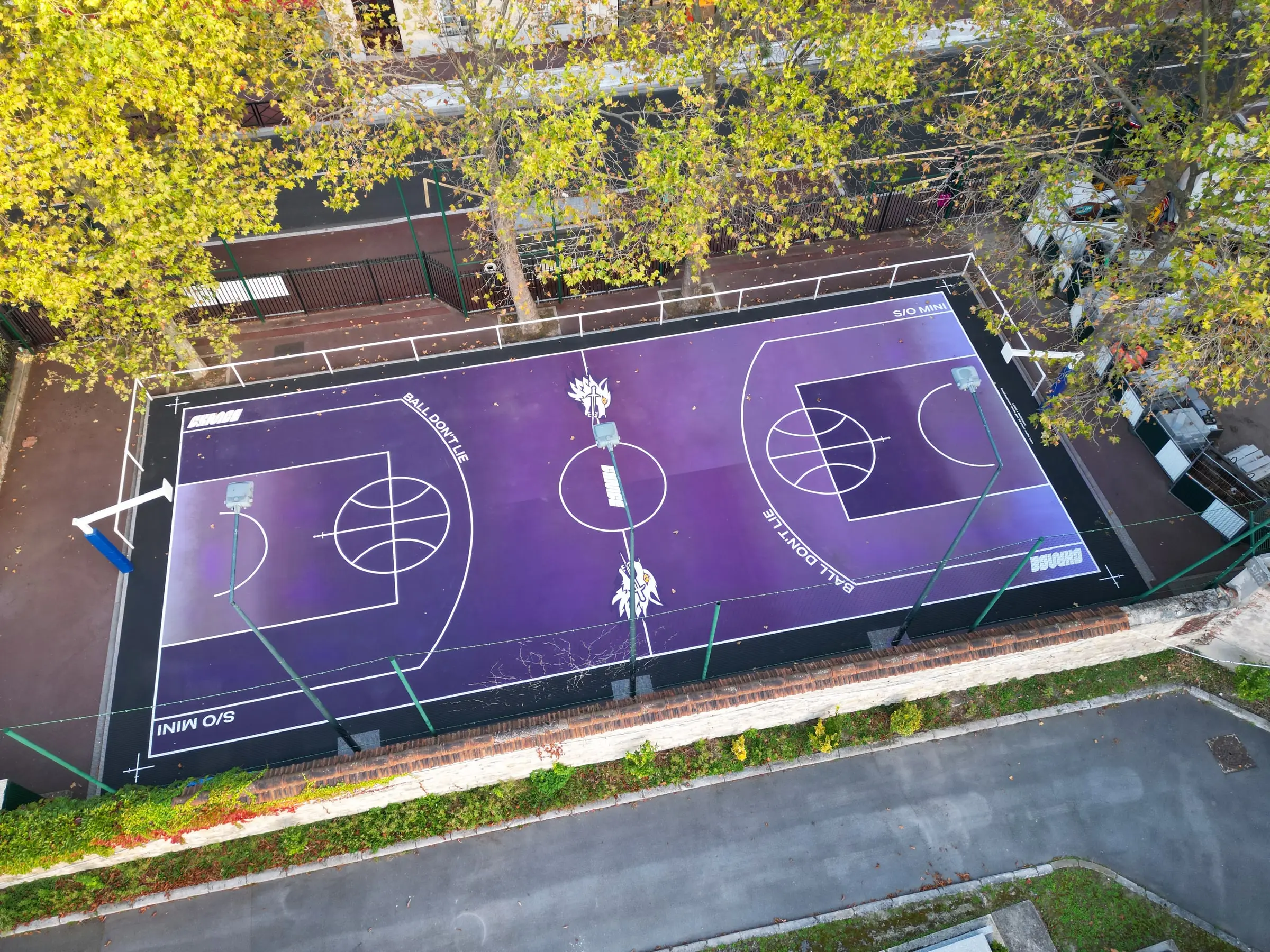 Vue aérienne d’un city-stade de basket peint en dégradé violet, entouré de platanes