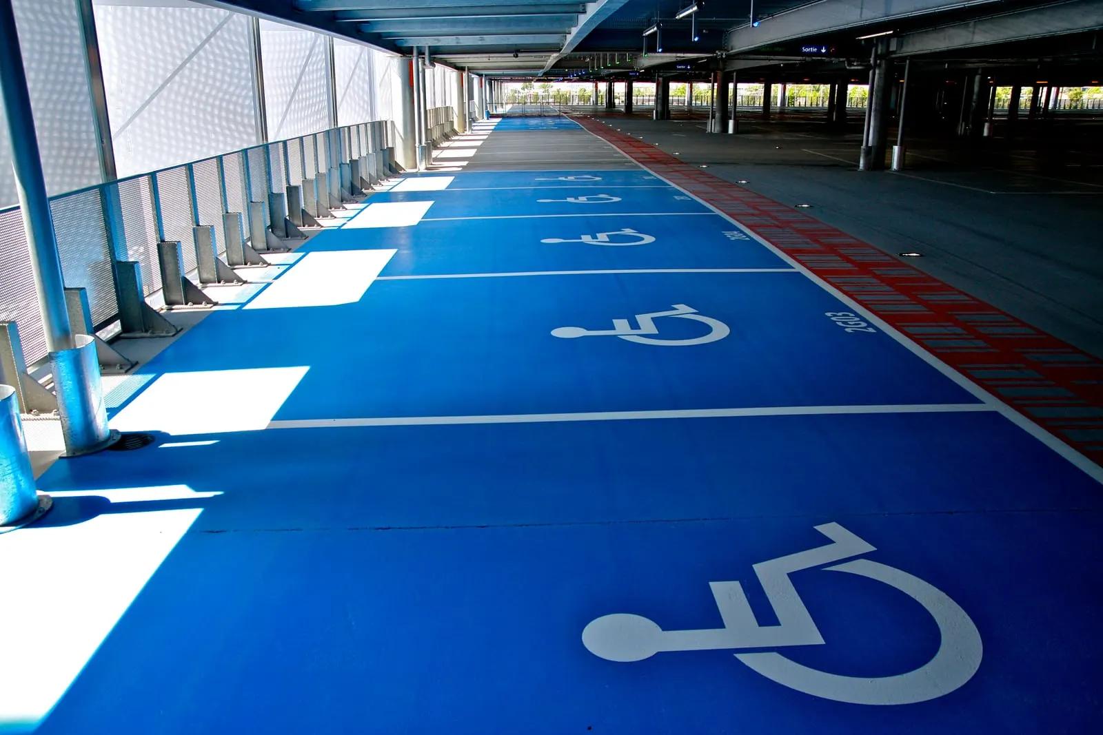 Rangée de places PMR à fond bleu et pictogramme, bordée du cheminement piéton rouge