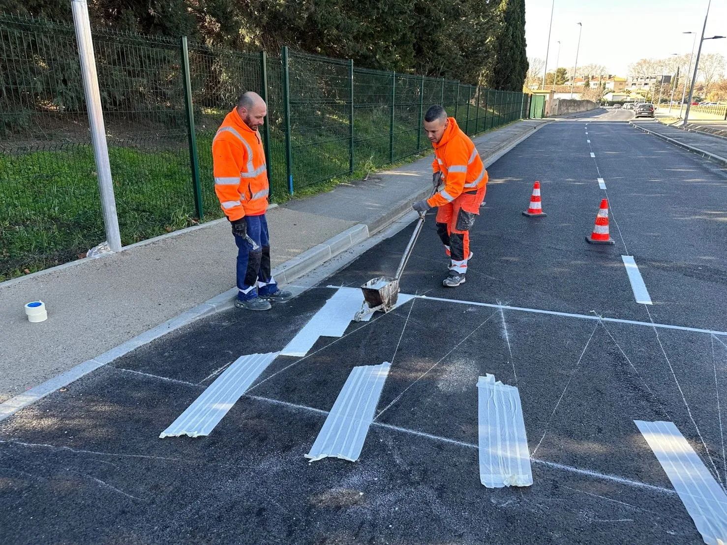Deux compagnons ASM traçant un passage piéton sur une voirie neuve, bandes protégées à l’adhésif et cônes de signalisation