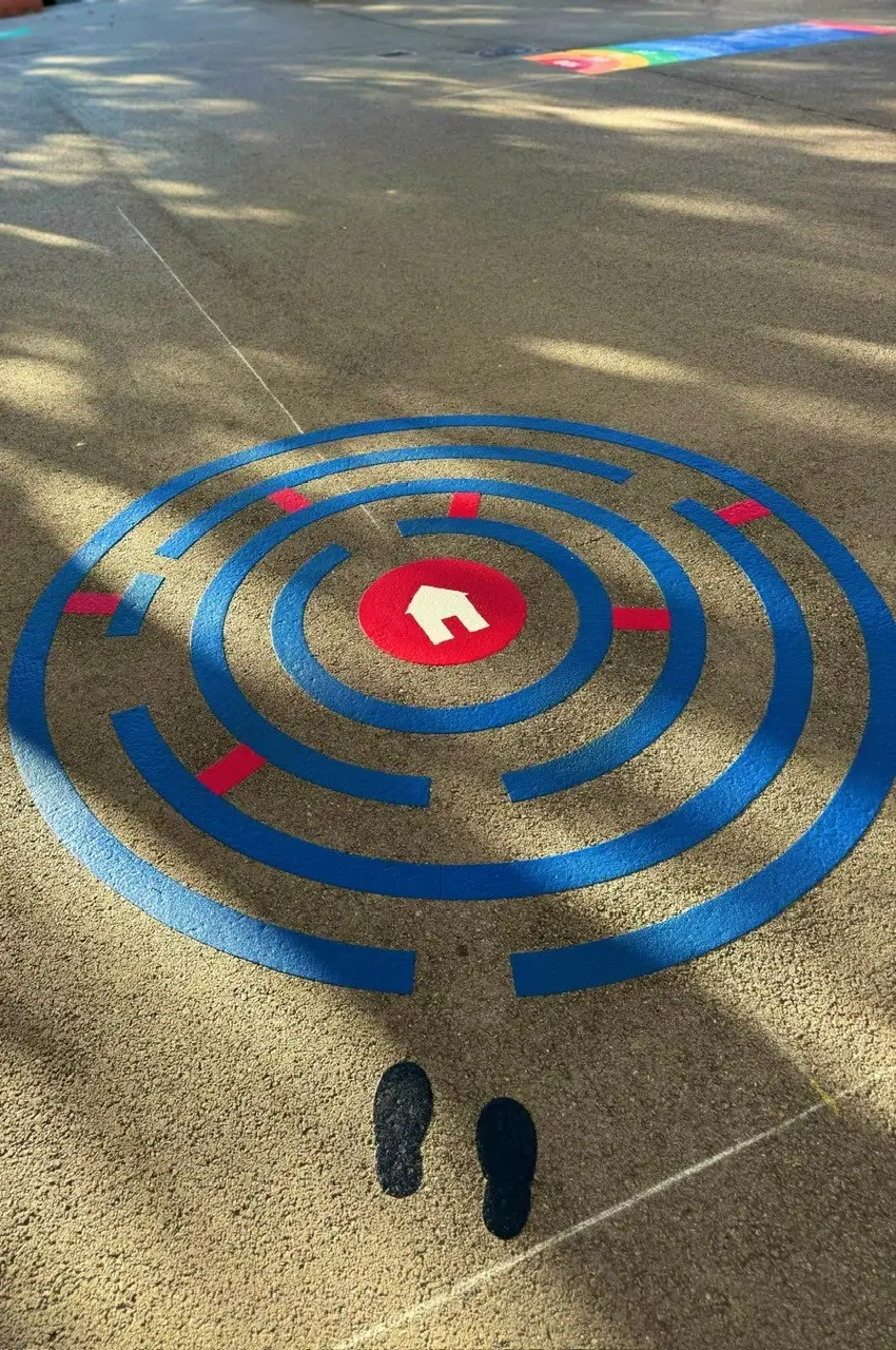 Labyrinthe circulaire bleu et rouge tracé au sol dans une cour d’école, empreintes de pas à l’entrée