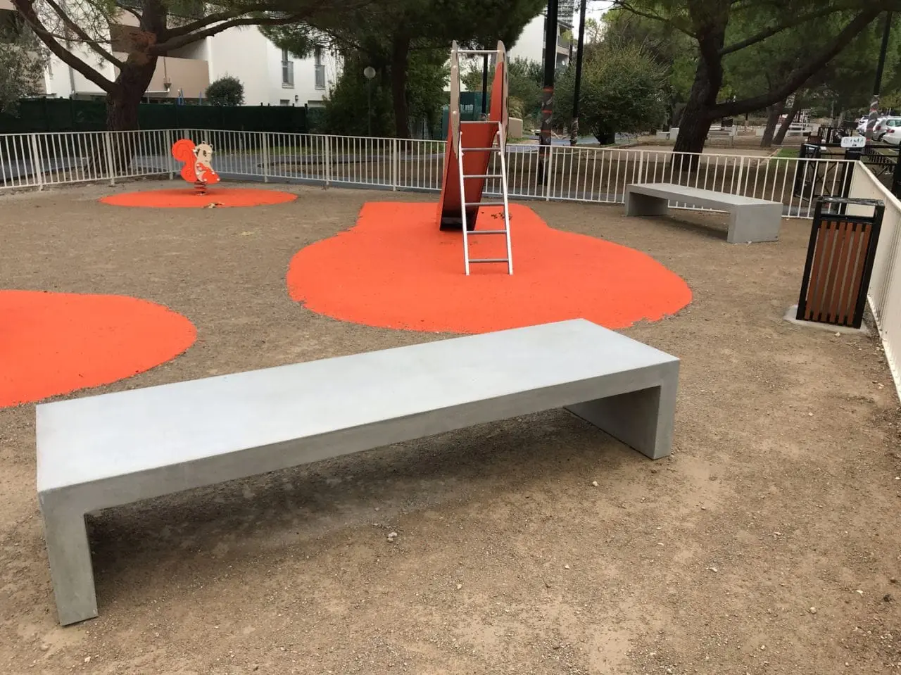 Banc béton clair posé au bord d’une aire de jeux au sol orange