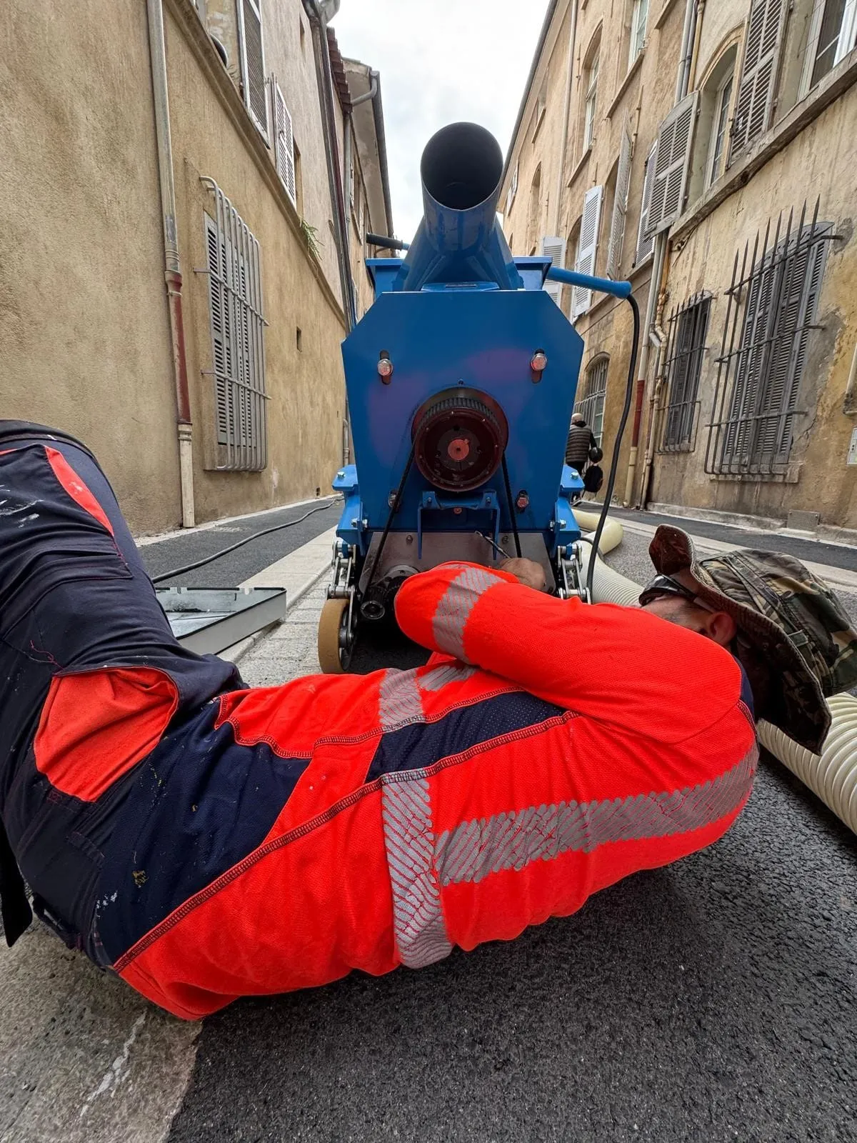Compagnon ASM allongé au sol dans une rue étroite, réglant la grenailleuse bleue avant le passage de préparation