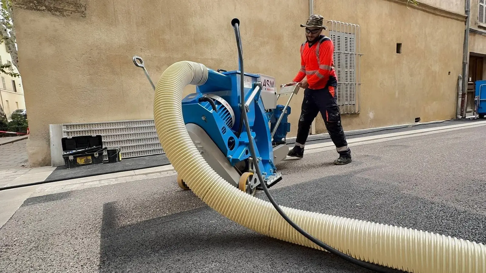 Compagnons ASM au grenaillage d’une rue, machine et flexible d’aspiration en pleine chaussée