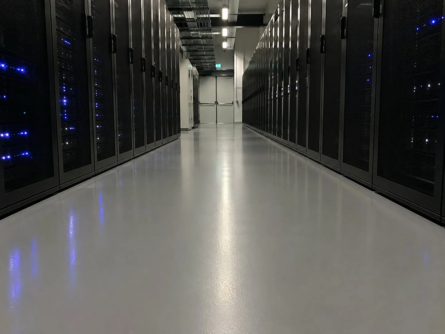 Allée de data center au sol résine gris clair très lisse, reflets des diodes bleues des baies de serveurs
