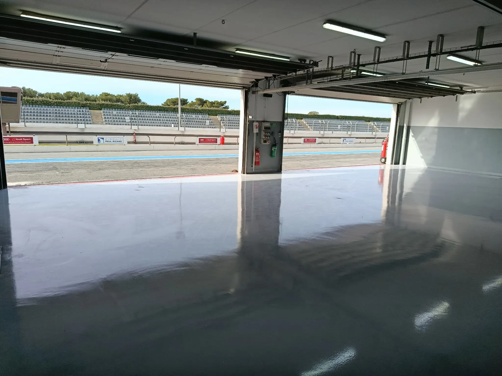 Sol résine gris brillant d’un box du Circuit Paul Ricard, portes ouvertes sur la voie des stands et les tribunes
