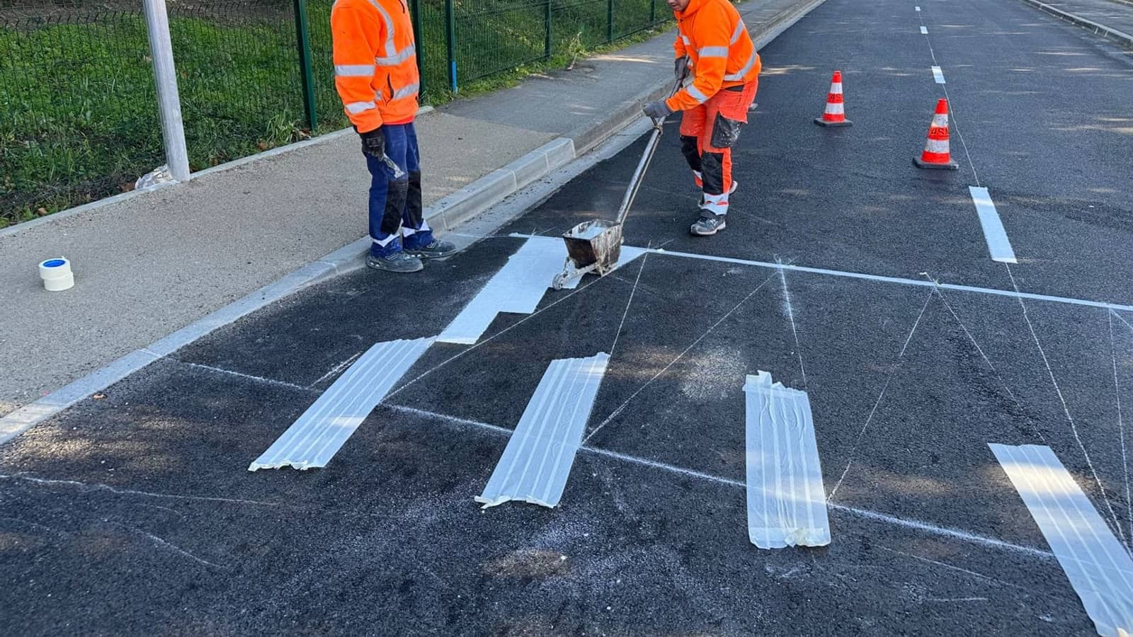 Deux compagnons ASM appliquant un marquage blanc de passage piéton sur une chaussée, cônes de signalisation à l'arrière-plan